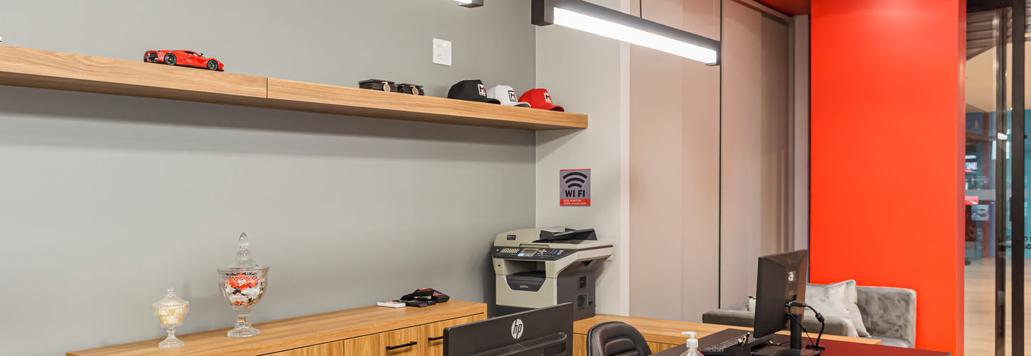 Detalhe da mesa de vendas com computadores, impressora, e cadeiras pretas. Foco na iluminação moderna e teto vermelho.