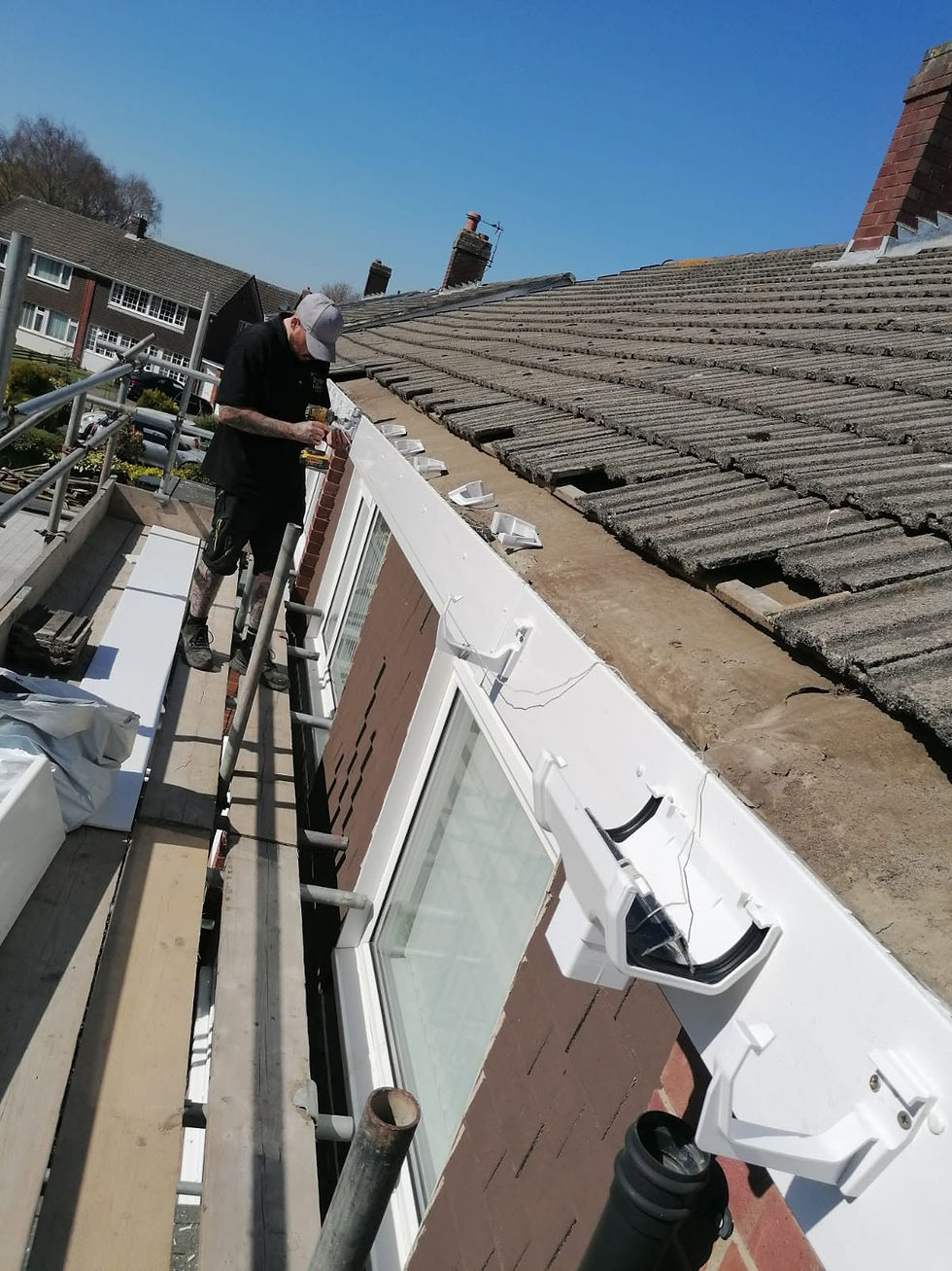 Worker installing white uPVC guttering on scaffolding beside a tiled roof