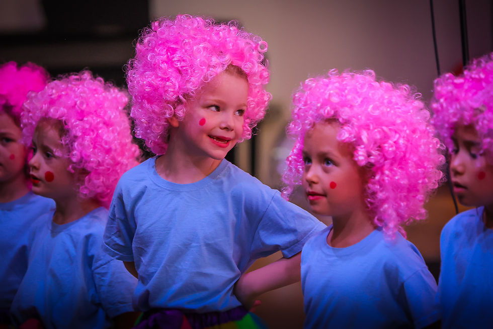 Young children in pink clown wigs and blue shirts performing on stage