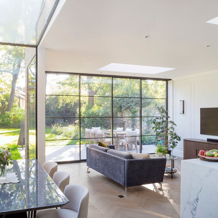 Sitting area, grey sofa with metal framed full height glazing beyond, on the left in the foreground is the edge of the dining area and on the right, the kitchen work surface