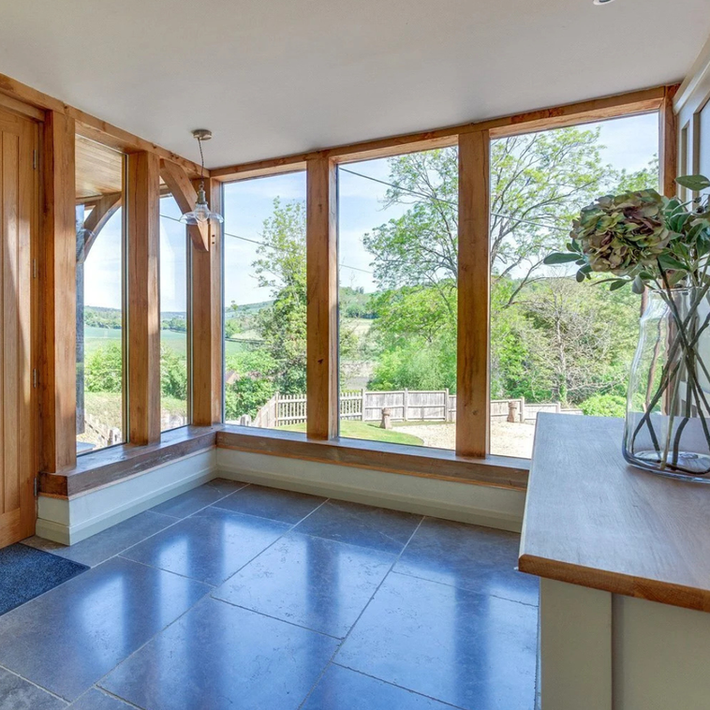 Light-filled entrance hall interior with floor-to-ceiling oak-framed windows overlooking countryside, built-in timber window bench seating, large format grey stone floor tiles, exposed oak structural posts and bracing, pendant lighting, and modern minimalist details including glass vase with hydrangeas.