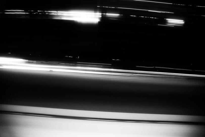 Travelling the light rail in Canberra, timelapse in the evening. The way to was full, couldn’t get a window seat. But heading back from the city is quieter, the murky windows diffuse the lights of the Northbourne corridor.  A black and white photograph by Canberra photographer Megan Kennedy, an abstract timelapse rendering of the busy city area through murky train windows