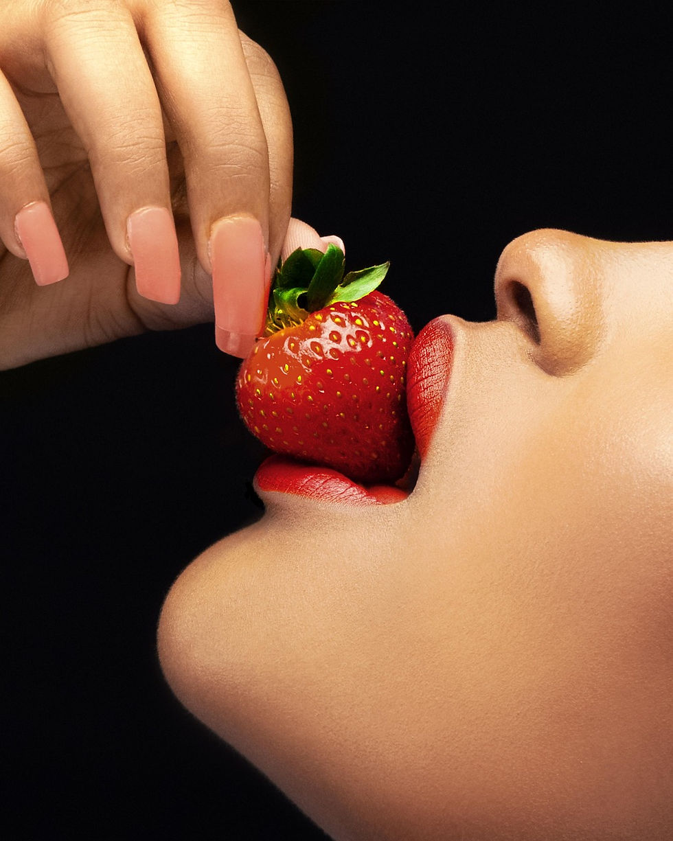 A woman's lips delicately embrace a ripe strawberry, highlighting the vibrant red hue against her smooth complexion.