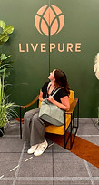 a young woman sitting in a chair infront of a backgrop that says Live Pure with a logo that looks like a 3 leaf lotus flower in a circle. She is looking back over her shoulder at the logo