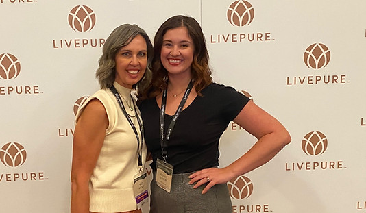 A mother, mid 50s in a white shirt and daughter mid 20s in a black shirt, standing together in front of a backdrops that says live pure in a step and repeat. There is a logo that looks like a lotus flower in a circle