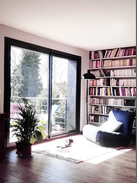 Coin lecture intérieur avec grande baie vitrée et lumière du soleil sur le parquet et la bibliothèque.