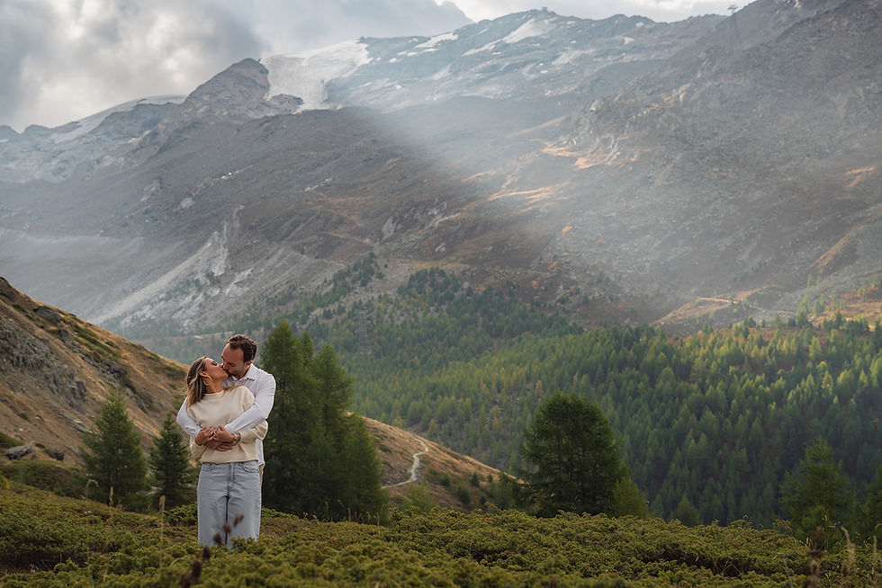 Couple embracing and kissing on a grassy mountain slope, with forested valley and snow-streaked peaks under soft light.
