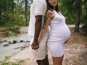 a couple holds hands back to back in gorgeous, rich colored forest. They stand alongside a stream as he closes his eyes in peaceful serenity and she holds her pregnant belly and smiles.
