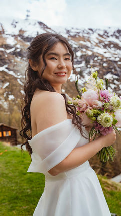 a beautiful young bride smiles happily in her gown with snow capped mountains and bright green grass behind her.