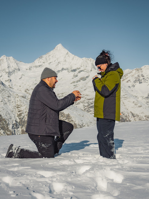 a man proposes to a woman in the snow with Swiss mountain peaks behind thems