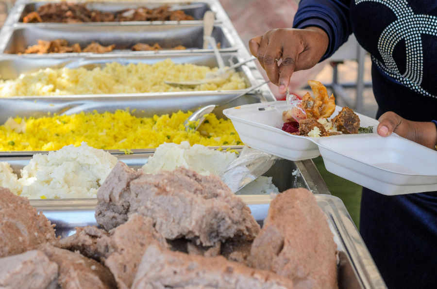 Person serving food onto a plate from a buffet with varied dishes, including rice and meat, in silver trays. Casual outdoor setting.