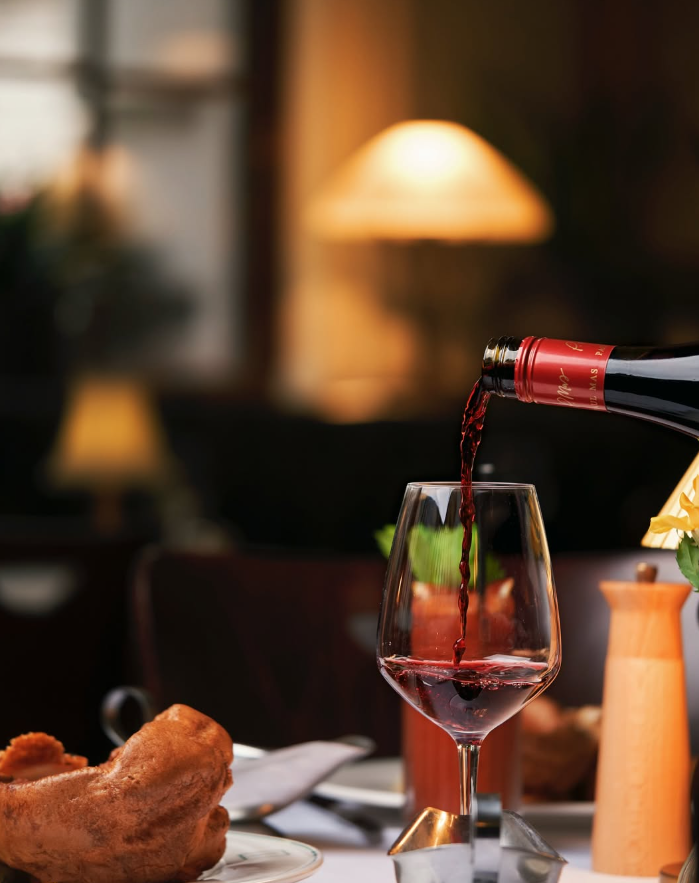 Red wine is poured into a glass on a dining table with warm lighting. Bread and a candle are visible, creating a cozy ambiance.