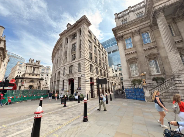 Historic buildings line a city street with people walking, including a woman in denim shorts. A red bus passes by, under a partly cloudy sky.