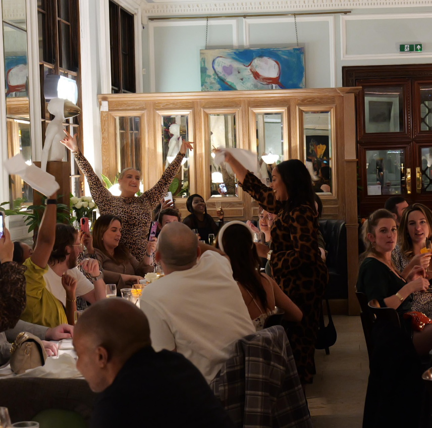 People celebrating at 1 Lombard Street restaurant, holding napkins up. Warm lighting, mirrors, and art in background. Joyful mood with lively energy.