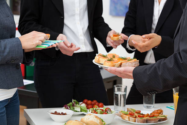 People in formal attire enjoy appetizers at a table, holding plates with food. The setting suggests a casual office gathering.