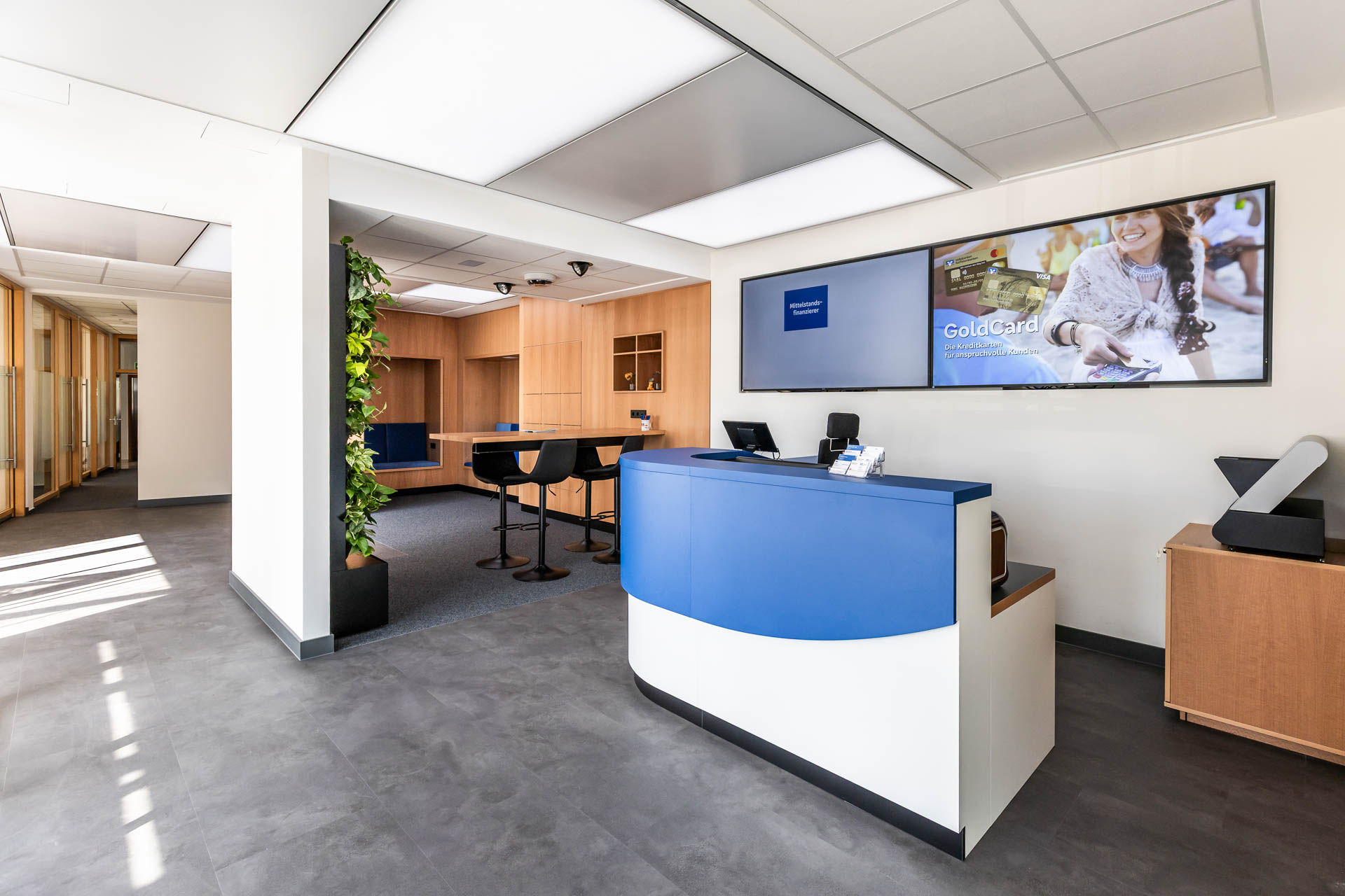 Blick in eine moderne Bankfiliale mit Sicht auf Infoscreens, einer grünen Wand und Bartisch