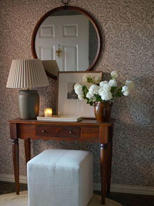 Console vintage en bois décorée de fleurs blanches, d’une lampe plissée, d’un cadre et d’un miroir rond devant un papier peint floral neutre signé Artza & Co.