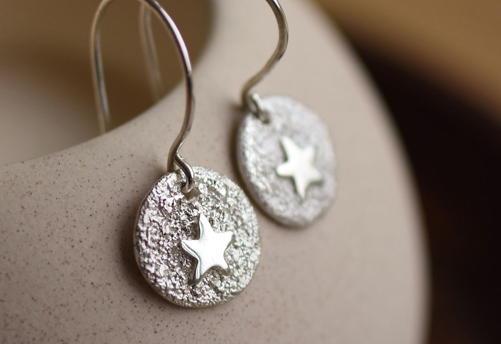 Thumbnail: silver round disc earrings with stars hanging from a beige ceramic dish