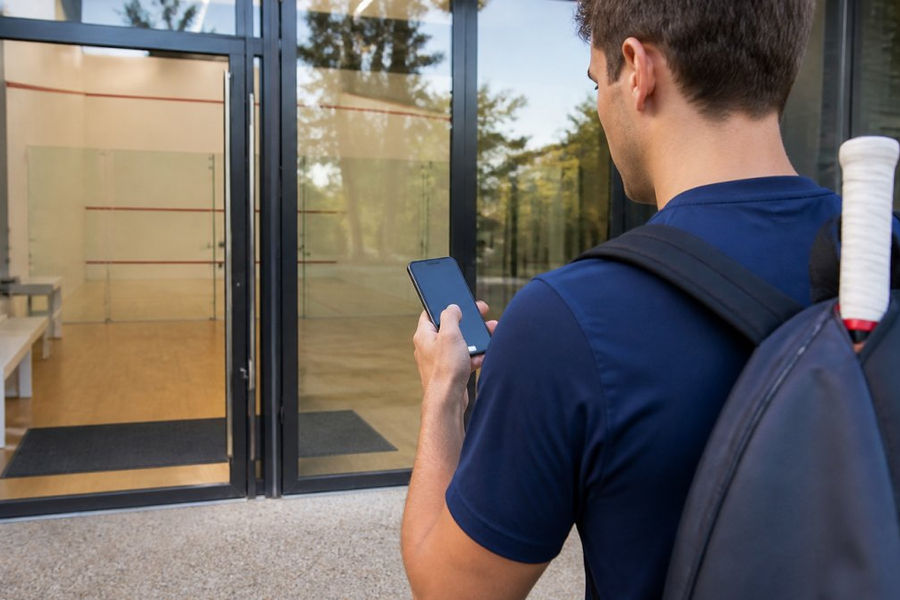 Player checking squash court hire rates on a smartphone app outside a modern sports facility entrance