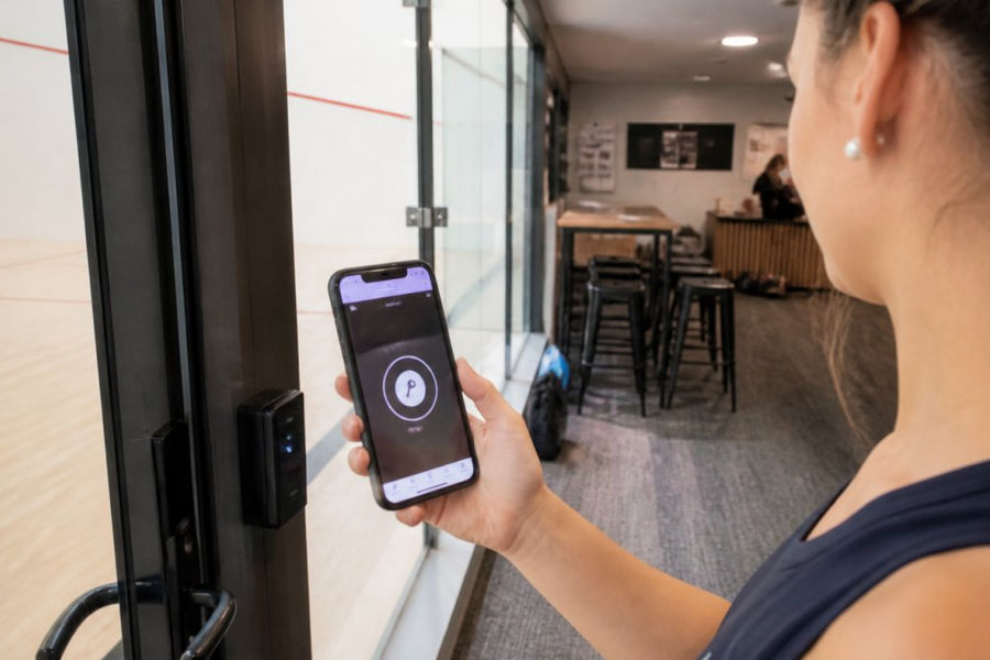 Player holding a smartphone displaying the SALTO app key interface, standing at the entrance of a modern squash court facility with the secure door access point visible in the background