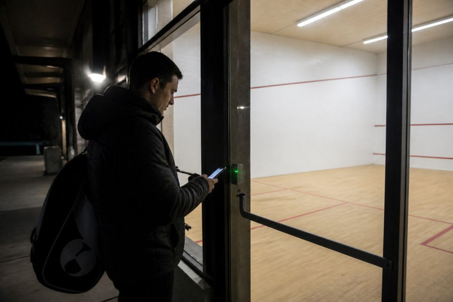 A squash player in a dark jacket standing at the glass door of a lit squash court at night, phone screen glowing as they unlock the entrance