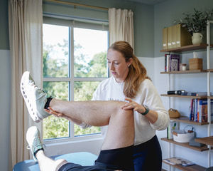 Bridget Sinha, Owner of Balanced Physical Therapy & Pilates in white checks patient's knee in a bright room with shelves and window view.