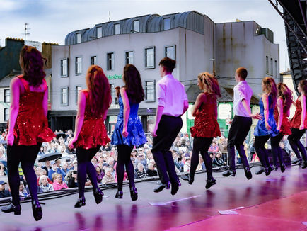 Irish Dancers in red and blue sparkly dresses and black pants perform onstage. A crowd watches from the street, with buildings as the backdrop.