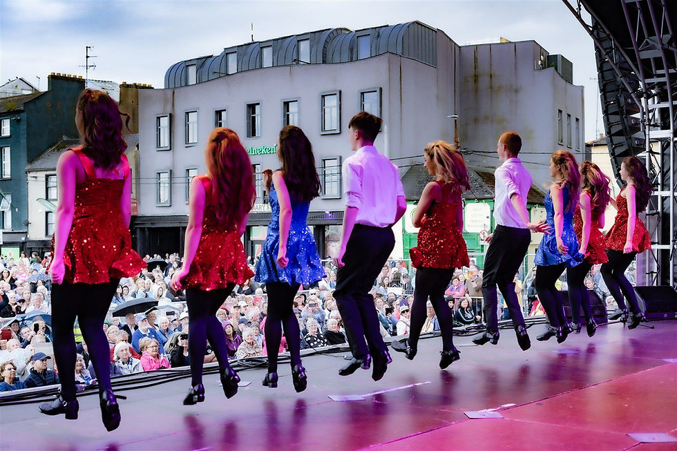 Irish Dancers in red and blue sparkly dresses and black pants perform onstage. A crowd watches from the street, with buildings as the backdrop.