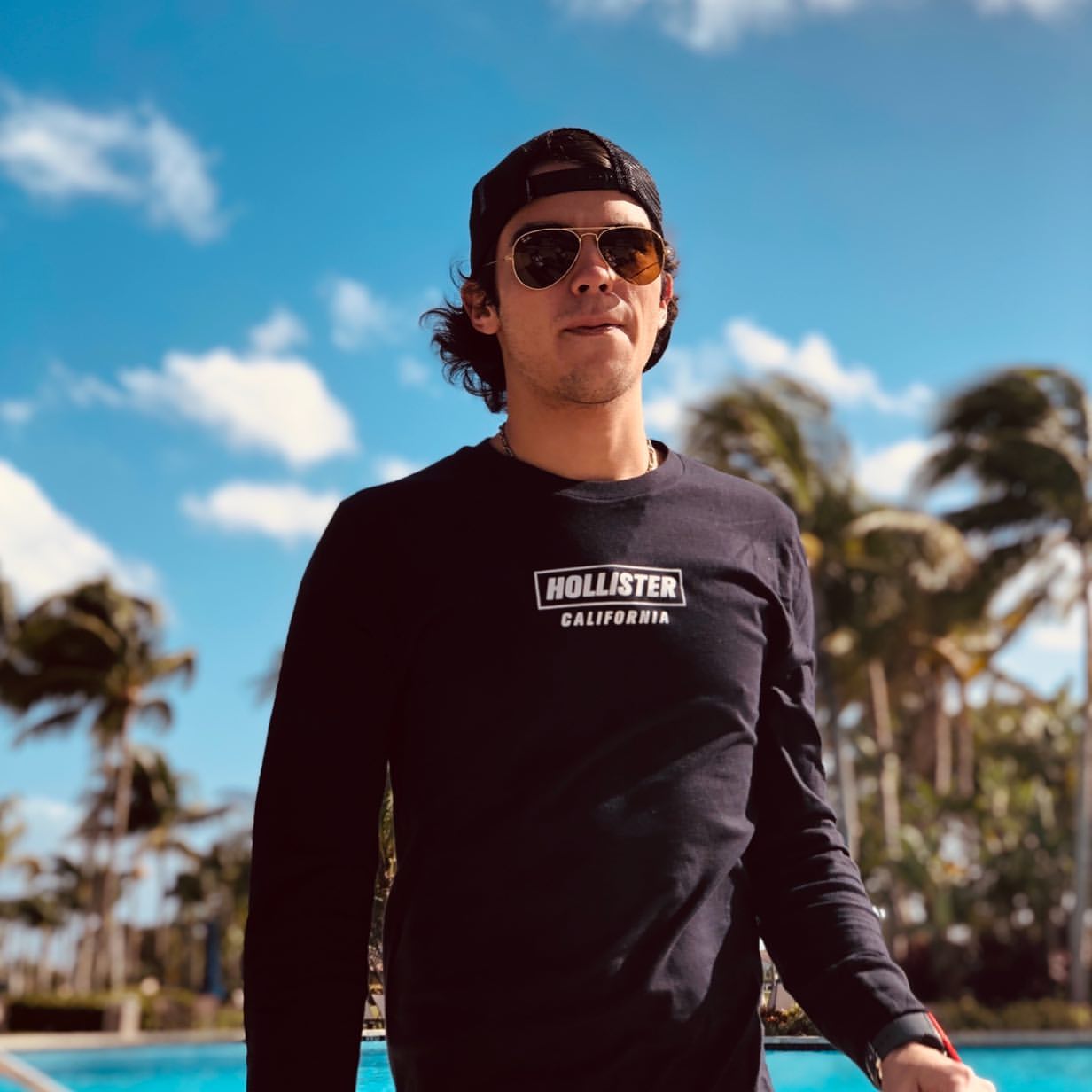 Hombre joven con gafas de sol y sudadera Hollister ante palmeras