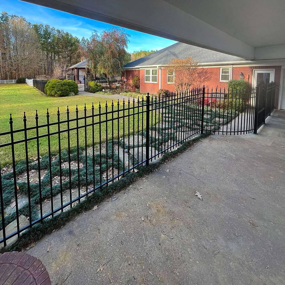Brick house with black iron fence, manicured lawn, and trees under a bright blue sky, seen from a shaded patio.