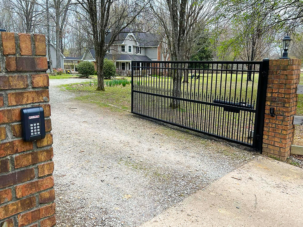 Open black driveway gate between brick pillars with keypad, leading to a gravel drive and house under leafless trees.