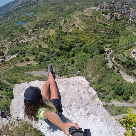 A woman sits on a large rock overlooking the mountainous village of Akoura, Lebanon, with a winding road cutting through the green landscape below.