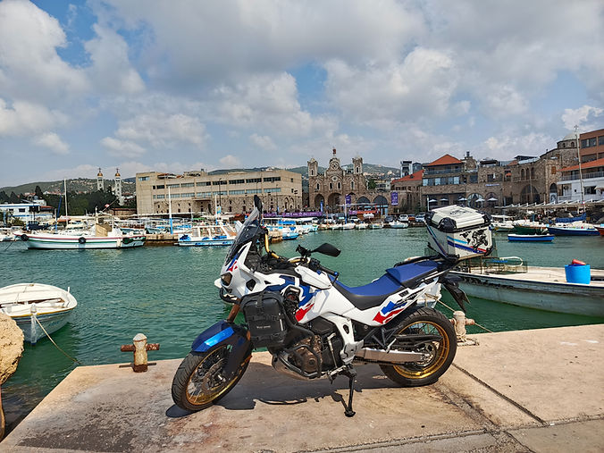 Honda Africa Twin parked at Batroun harbor with fishing boats and the old stone waterfront in the background.