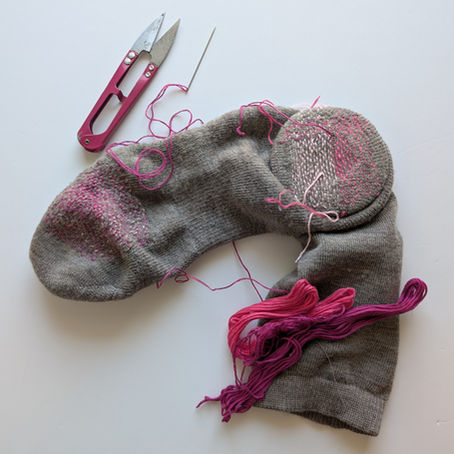 a sock with darning on the toe and heel lies next to pink thread snips, a needle with pink thread, and various shades of pink skeins of embroidery thread on a white background
