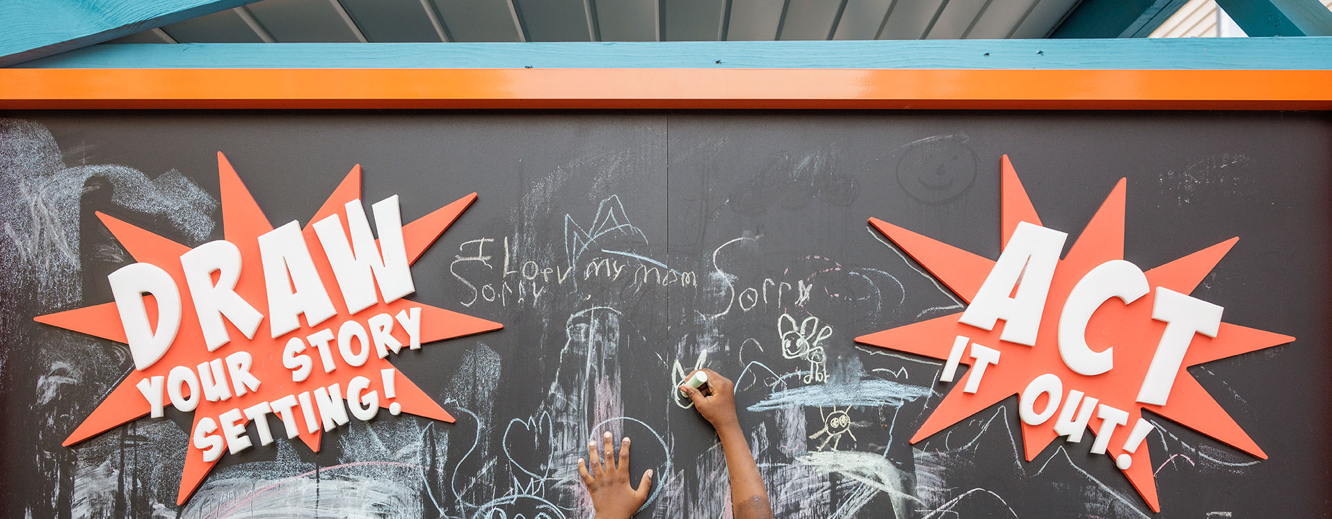 Little boy draws on a big chalkboard surrounded by a prompt "Draw your story setting!"