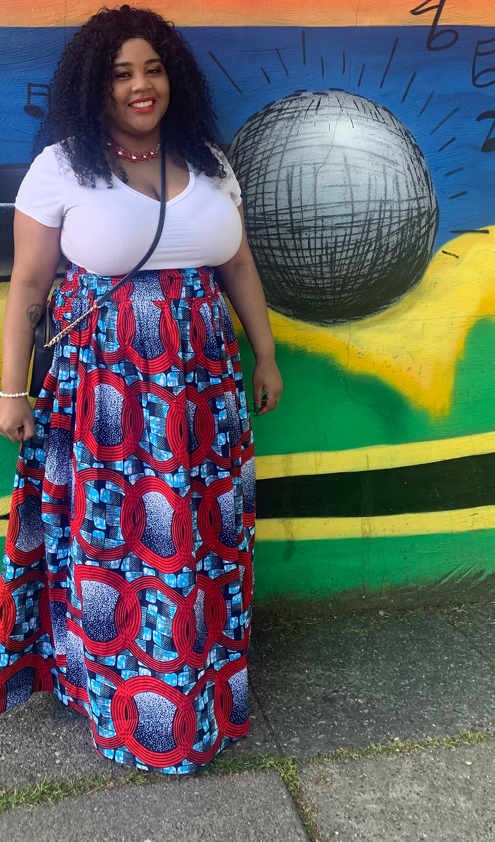 A woman with a white shirt and red skirt smiling behind a mural.