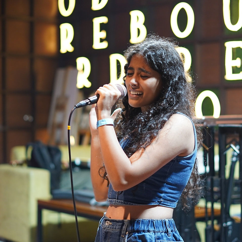 Vidyanshi Rajwadha sings into a microphone on a DHE Studio stage, eyes closed, with glowing letters and warm lights behind her.
