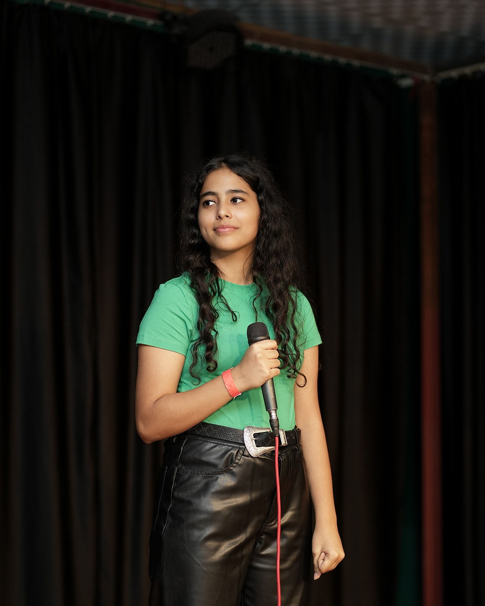 Vidyanshi Rajwadha in a green shirt and black pants holds a microphone on a dark stage, looking calm against black curtains.