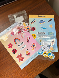 Two different customized sticker sheets for two kids. One pink with a small baggie of smaller loose stickers and a blue sheet with a bag of  smaller loose stickers.