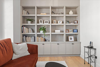 Living room featuring built-in gray bookshelves filled with books, framed photos, decor items, and plants. A rust-colored sofa with a textured white pillow is in the foreground, while a small black side table with a phone is placed to the right