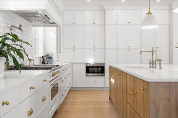 Modern kitchen with a mix of white and wood finishes, featuring a white cabinetry wall with gold hardware, a sleek built-in oven, and a wooden island with a white countertop. Decorative elements include gold-accented fixtures, potted plants, and a gas stove with a metallic backsplash faucet.