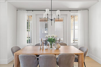 Bright dining room with a natural wood table surrounded by gray upholstered chairs. A modern black chandelier with white shades hangs above the table, which is decorated with a vase of white roses and minimalist decor. White curtains frame large windows, offering a view of the outdoor patio