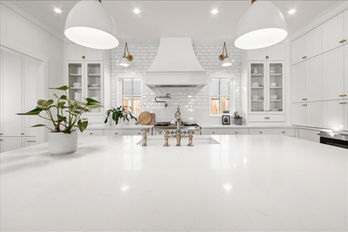 Minimalist kitchen featuring a spacious white island with a sleek gold faucet, surrounded by white cabinetry and a subway tile backsplash. The decor includes green plants, brass pendant lights, and a wooden cutting board for a touch of warmth.