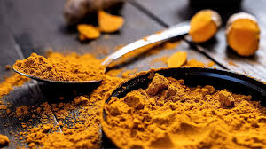 A black bowl and spoon filled with vibrant yellow turmeric powder on a dark wooden surface. Turmeric roots are blurred in the background.