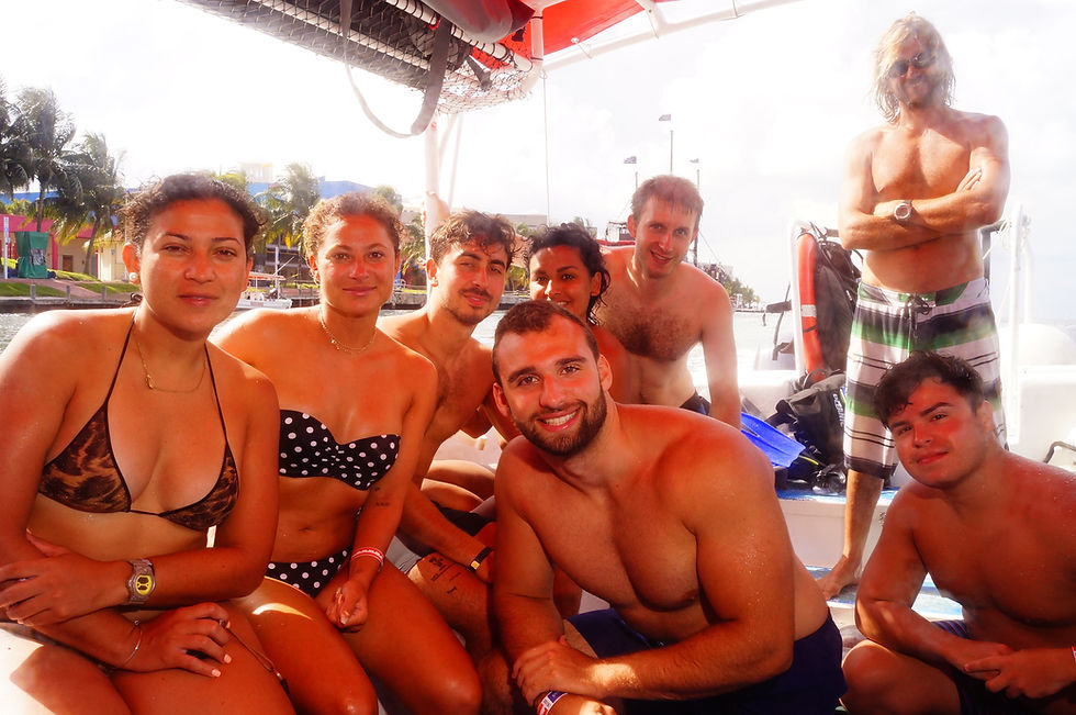Eight smiling people in swimsuits gathered on a boat under a red canopy, with colorful buildings and palm trees in the background.