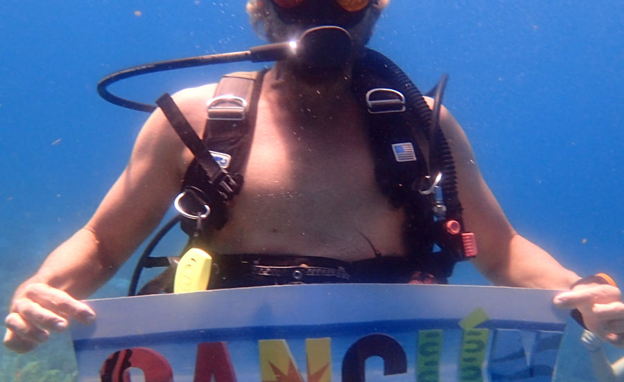 A scuba diver underwater holds a colorful "CANCUN" sign. They have blond hair and wear a diving mask. Blue ocean background.