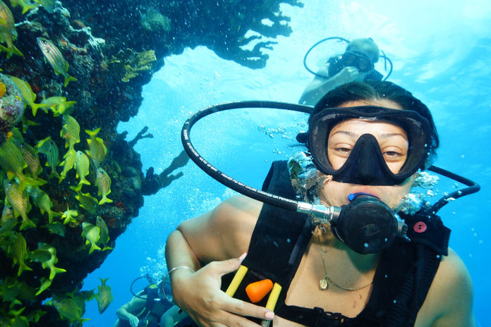 Scuba diver swims near coral reef with colorful fish in clear blue water, wearing a black mask and tank, exuding a sense of adventure while scuba diving Cancun.