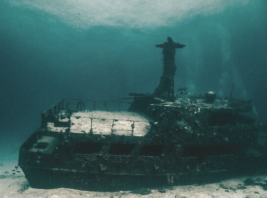 Sunken shipwreck underwater, surrounded by blue-green hues, with a solitary figurehead atop. Silty ocean floor visible, creating an eerie mood.
