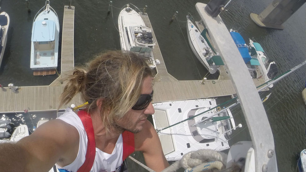Man in a harness ascends a boat mast, wearing sunglasses, with moored boats and a bridge in the background. Calm, focused expression.