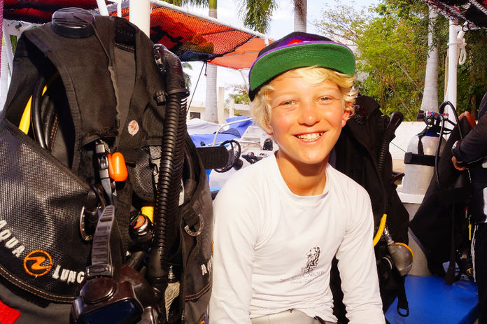 Young person smiling on a sunny patio, seated next to scuba gear with visible “Aqua Lung” text. Background shows tropical plants, ready for scuba diving Cancun.